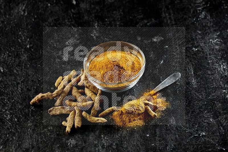 A glass bowl and a metal spoon full of turmeric powder and dried whole fingers next of them on textured black flooring