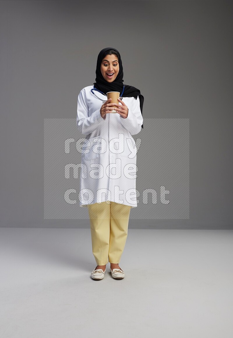 Saudi woman wearing lab coat with stethoscope standing holding paper cup on Gray background