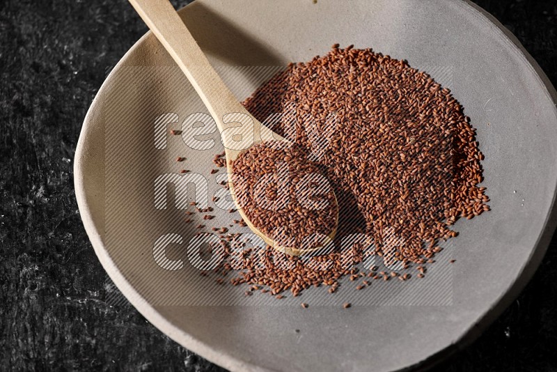 A multicolored pottery plate full of garden cress seeds and wooden spoon full of seeds on a textured black flooring