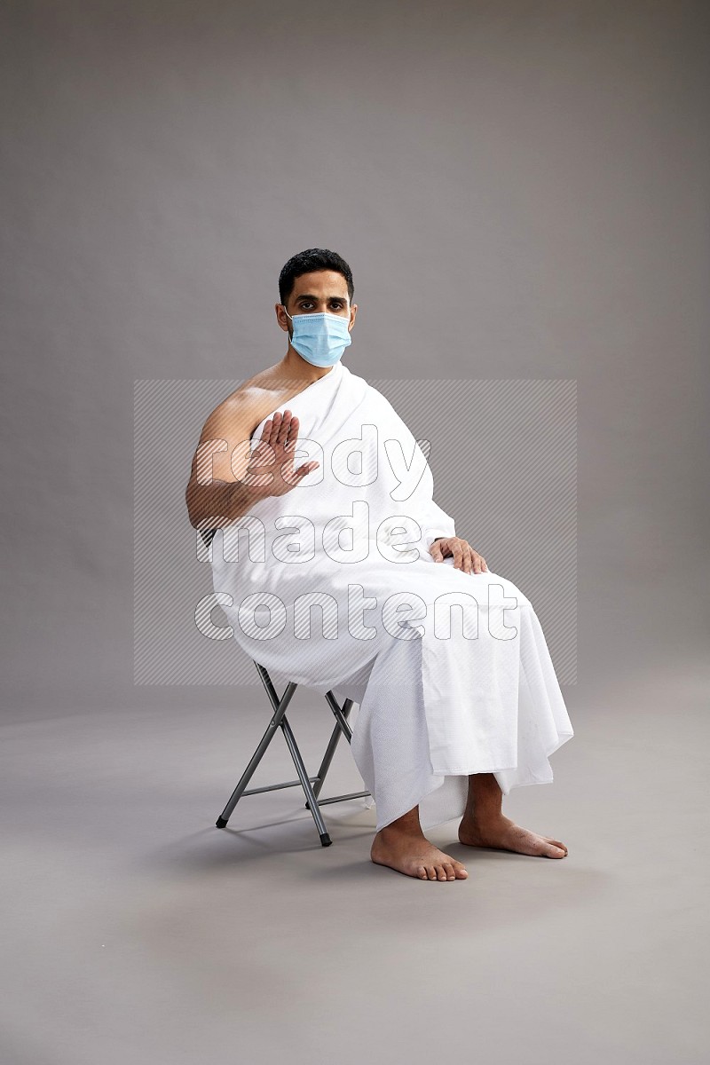 A man wearing Ehram with face mask sitting on chair Interacting with the camera on gray background