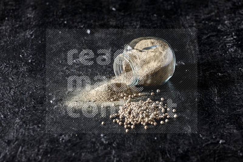 A flipped herbal glass jar full of white pepper powder with spilled powder and pepper beads on textured black flooring