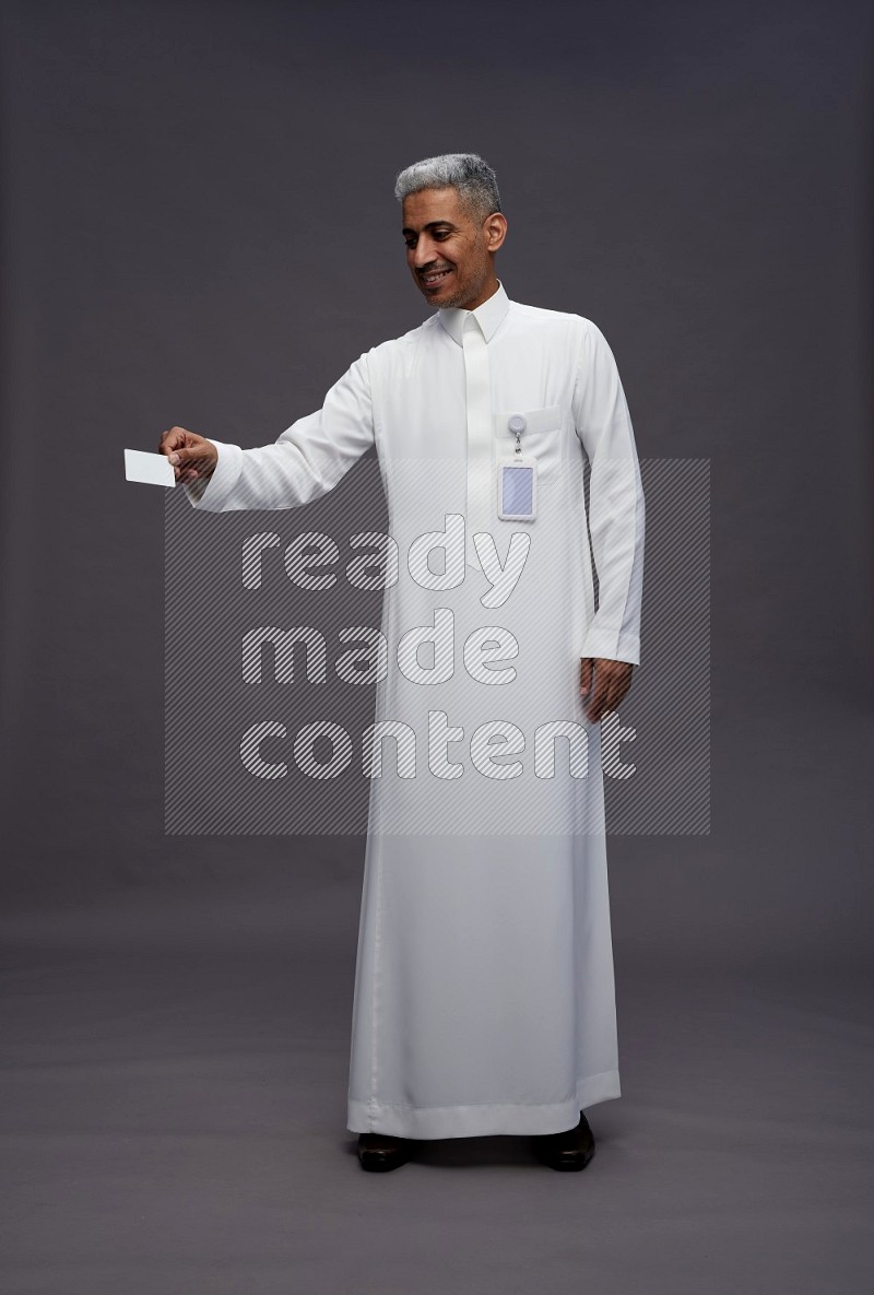 Saudi man wearing thob with pocket employee badge standing holding ATM card on gray background