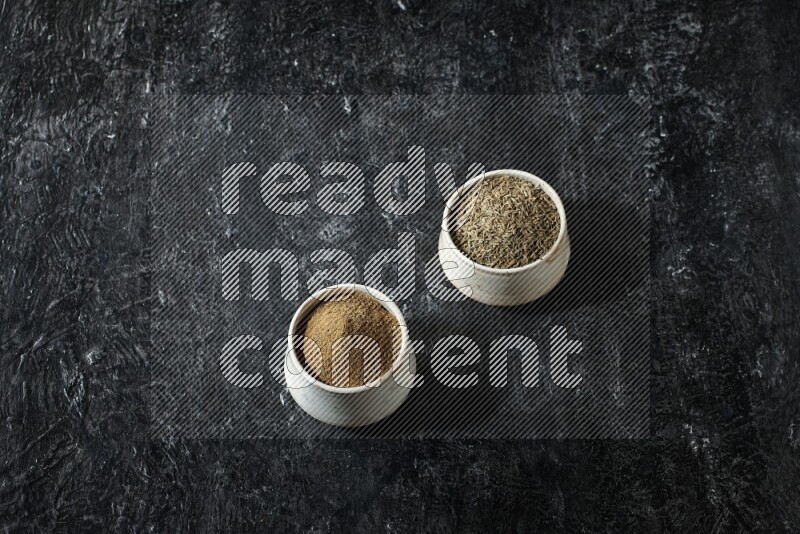 2 beige bowls, one full of cumin powder and the other full of seeds on a textured black flooring
