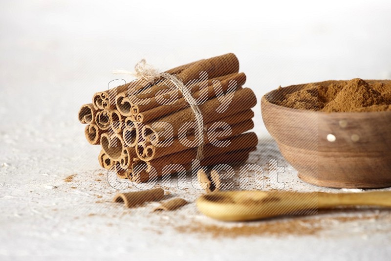 Cinnamon sticks stacked and bounded beside a wooden bowl full of cinnamon powder and a wooden spoon full of powder on white background