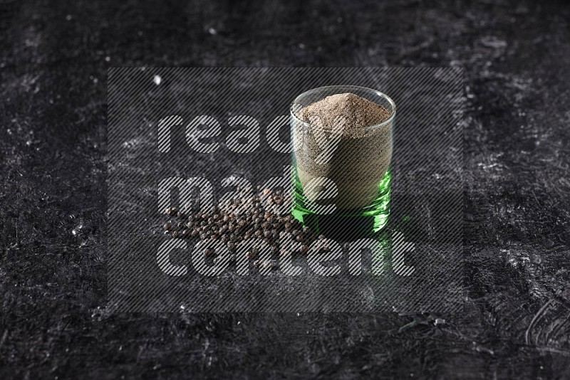 A glass cup full of black pepper powder with beads beside it on a textured black flooring