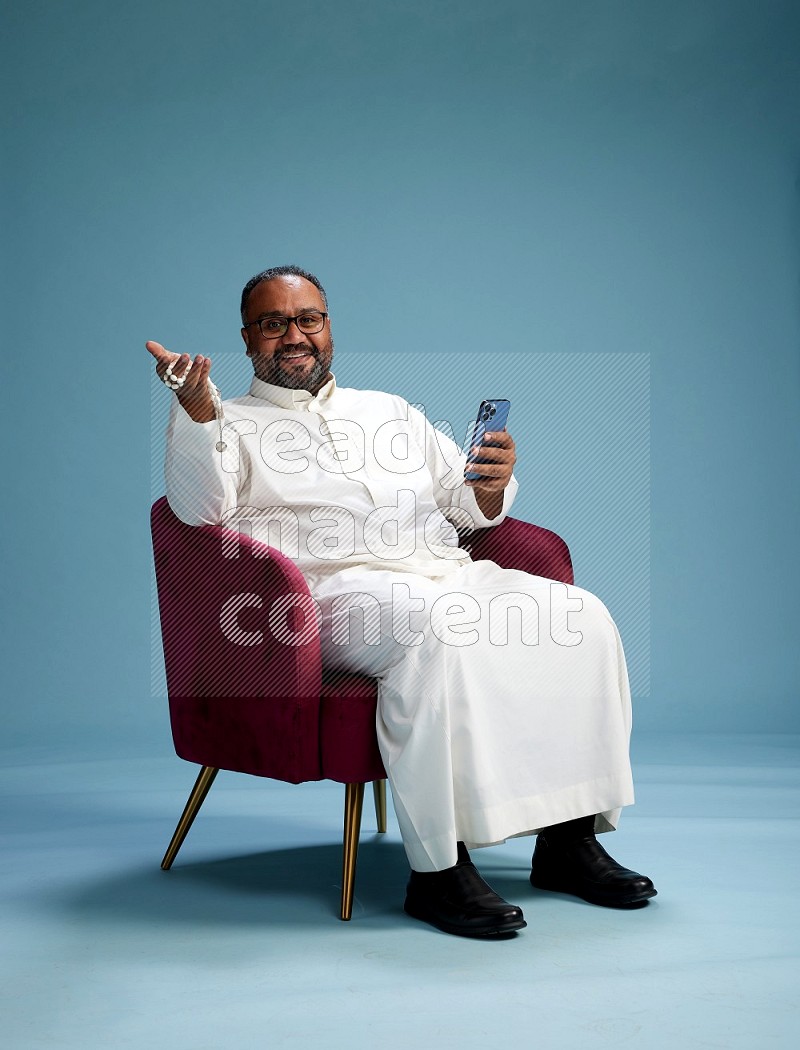 Saudi Man without shimag sitting on chair texting on phone on blue background