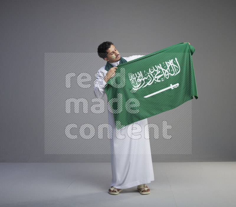 A Saudi man standing wearing thob with face painting wrapping Saudi flag scarf and holding big Saudi flag on gray background