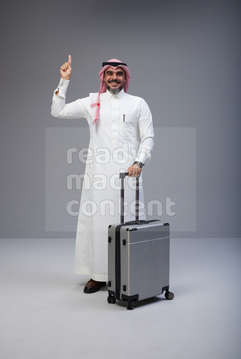 Saudi man wearing Thob and red Shomag standing holding Travel bag on Gray background