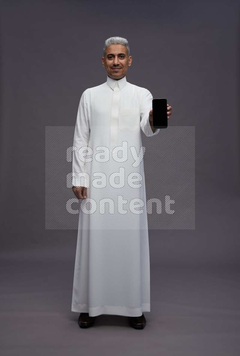 Saudi man wearing thob standing showing phone to camera on gray background