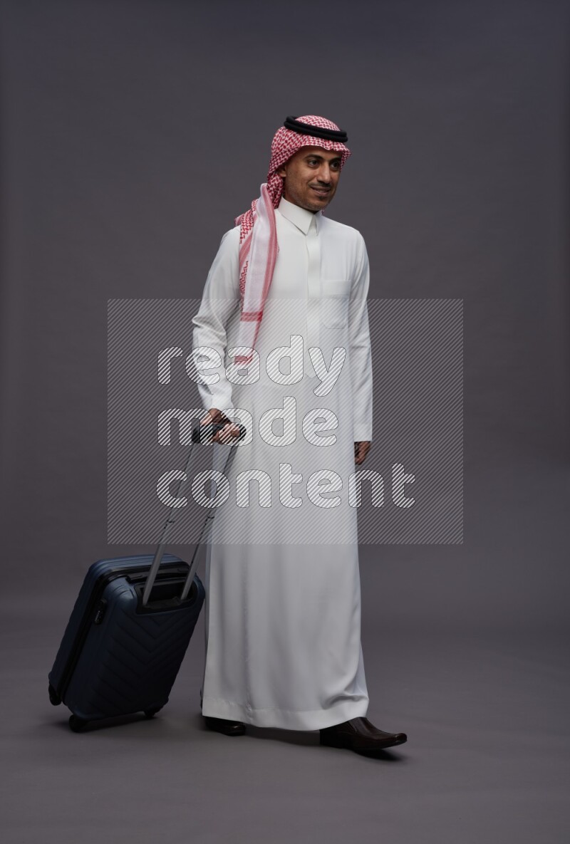 Saudi man wearing thob and shomag standing holding bag on gray background