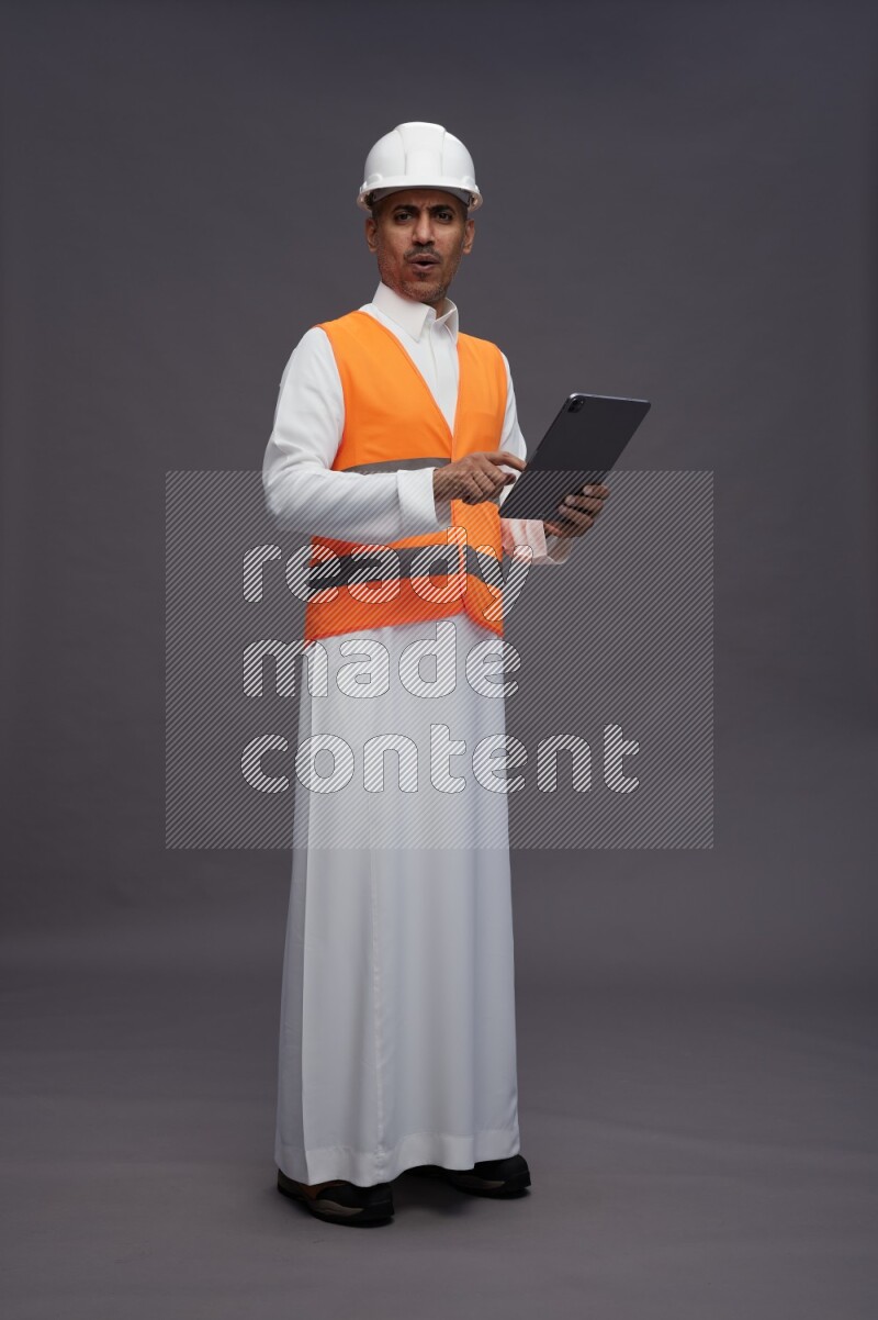 Saudi man wearing thob with engineer vest standing working on tablet on gray background