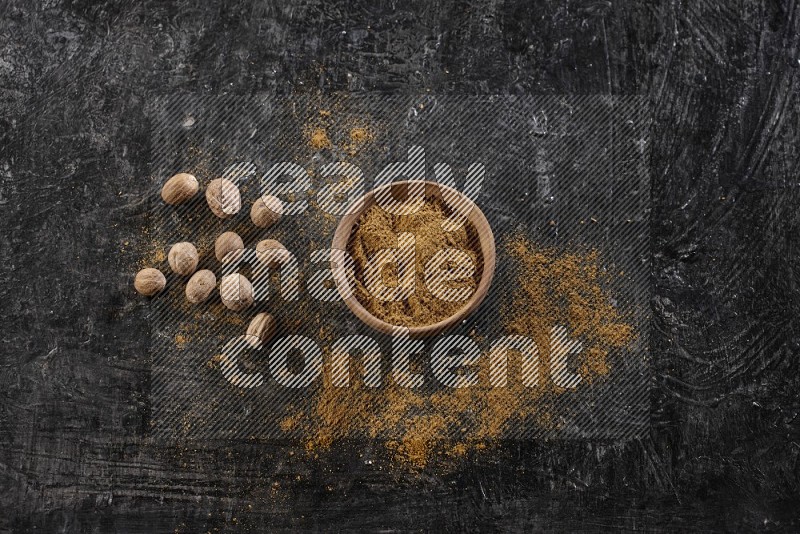 A wooden bowl full of nutmeg powder with whole seeds and sprinkled powder beside it on a textured black flooring