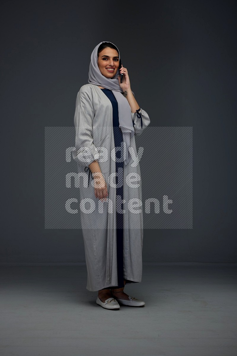 A Saudi woman wearing a light gray Abaya and head scarf standing and talking in the phone eye level on a grey background