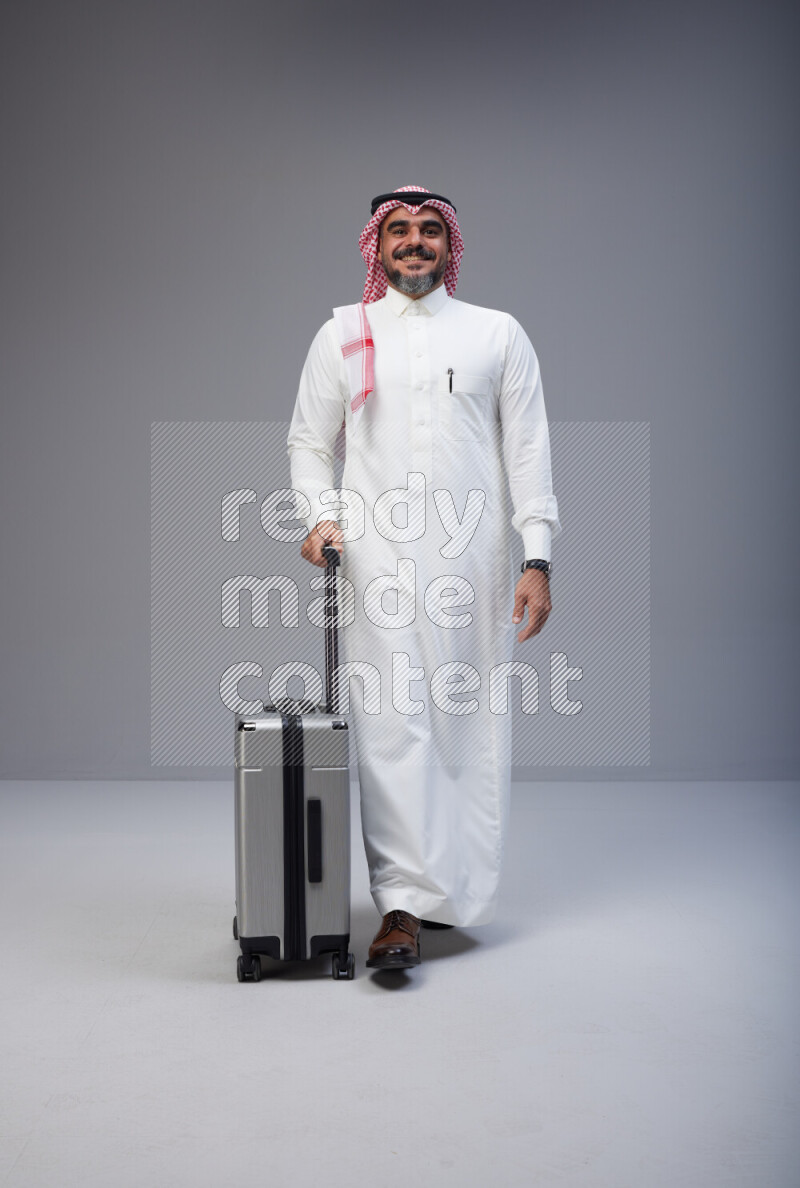 Saudi man wearing Thob and red Shomag standing holding Travel bag on Gray background