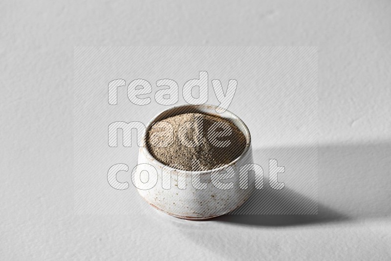 A white ceramic bowl full of black pepper powder and black pepper beads spread on white flooring