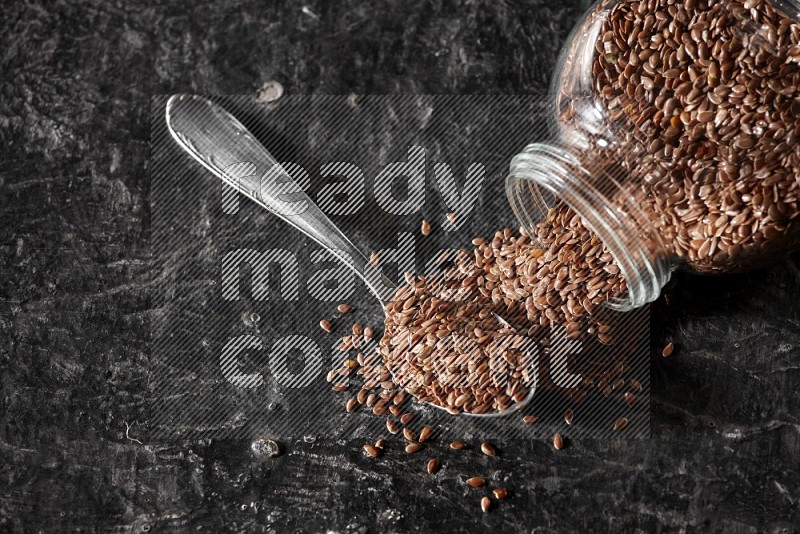 A glass spice jar full of flax flipped and seeds spread out with a metal spoon full of the seeds on a textured black flooring in different angles