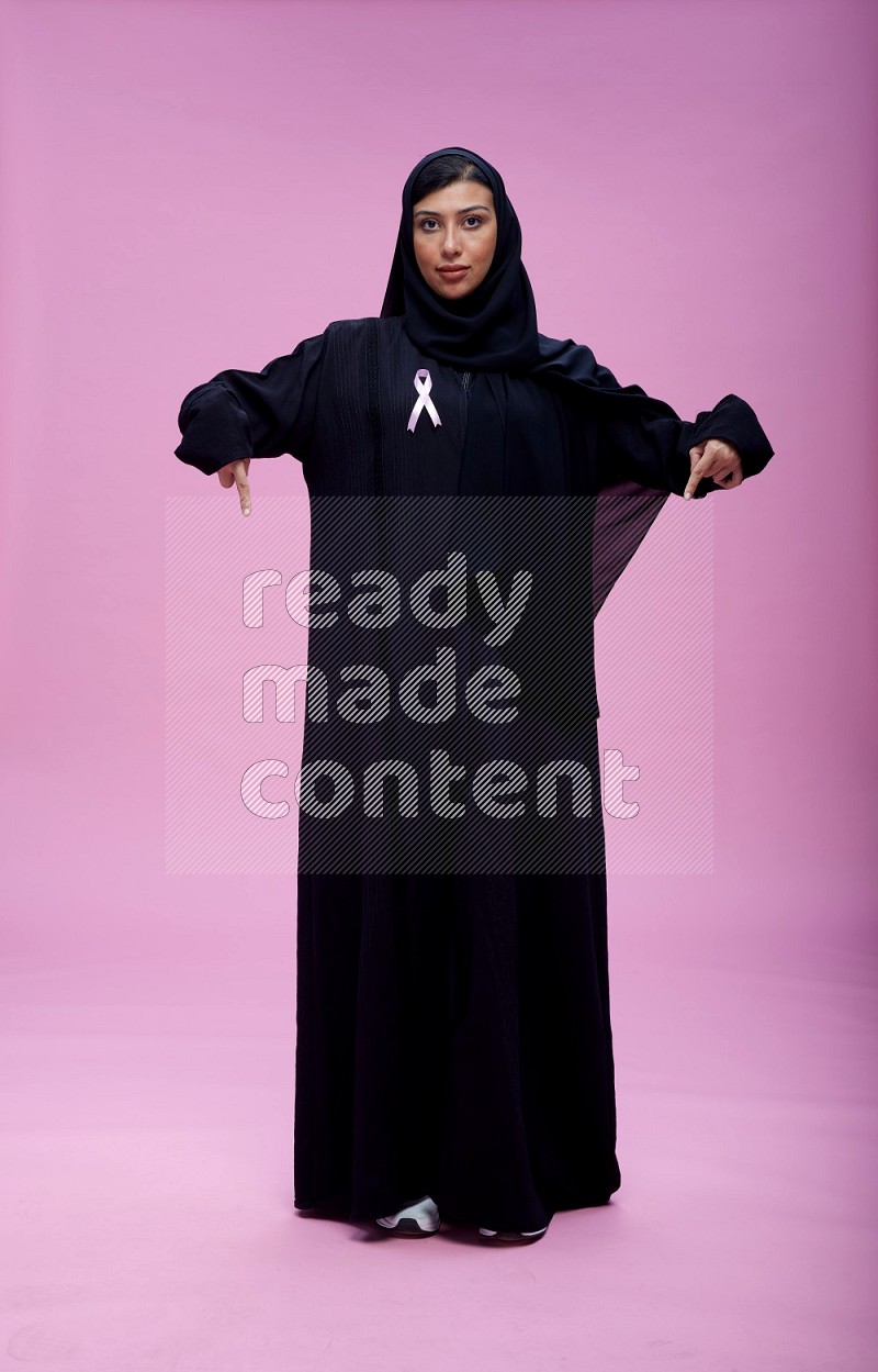 Saudi woman wearing pink ribbon on Abaya standing interacting with the camera on pink background