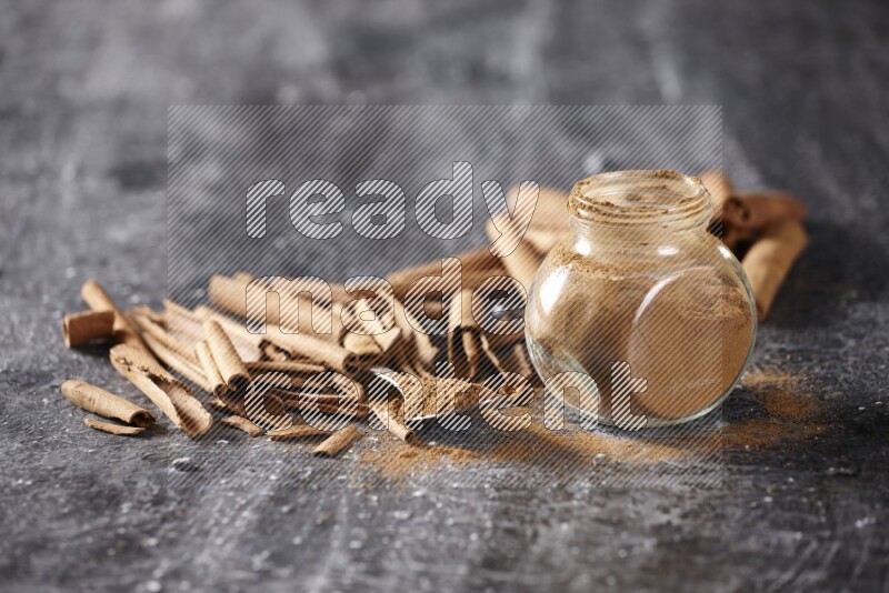 Herbal glass jar and a metal spoon full of cinnamon powder surrounded by cinnamon sticks on textured black background