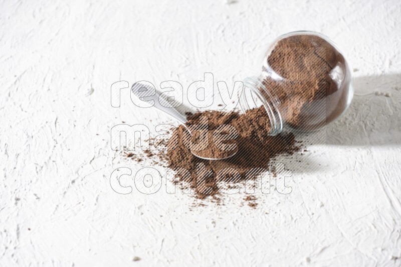 A flipped glass spice jar and a metal spoon full of cloves powder and powder came out of the jar on textured white flooring