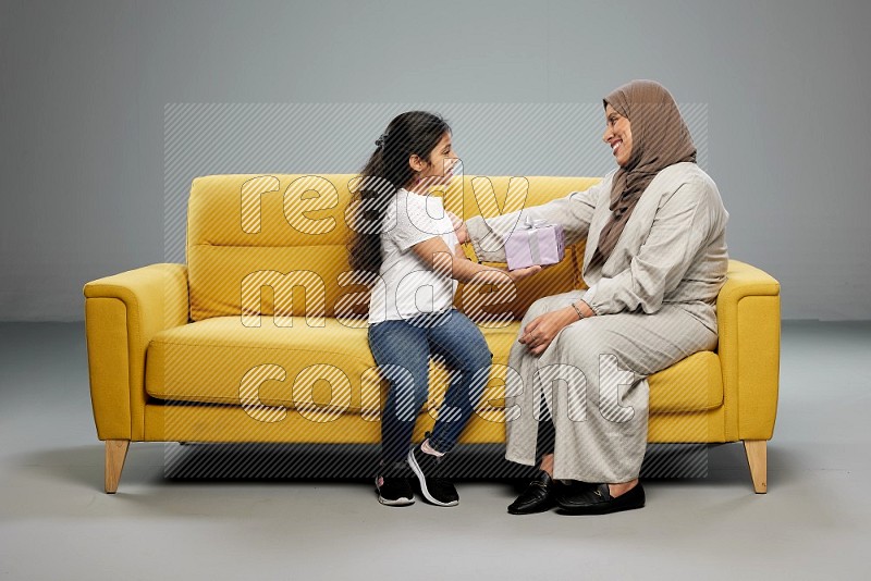 A girl sitting on a yellow sofa and giving a gift to her mother on gray background