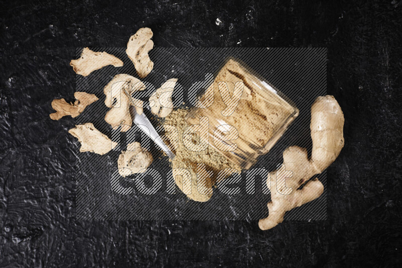 A glass jar full of ground ginger powder flipped with some spilling powder on black background