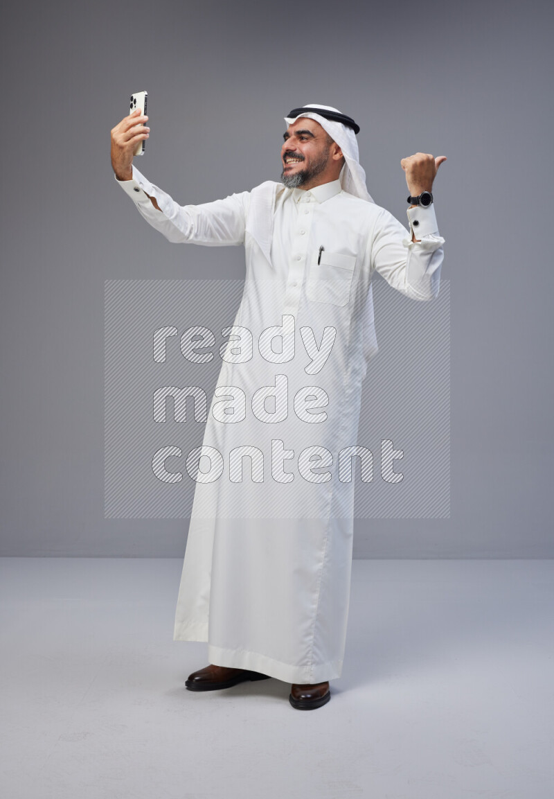 Saudi man Wearing Thob and white Shomag standing taking selfie on Gray background