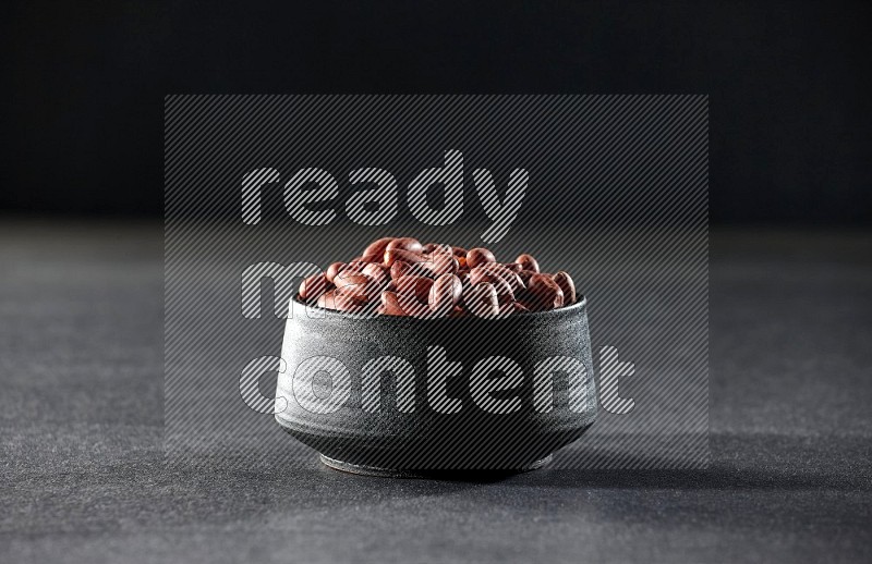 A black pottery bowl full of red skin peanuts on a black background in different angles