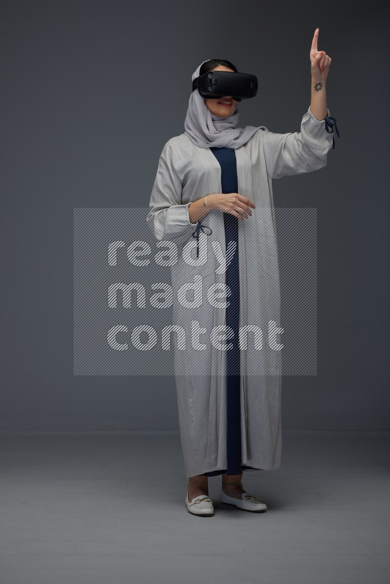 A Saudi woman wearing a light gray Abaya and head scarf standing and wearing VR eye level on a grey background