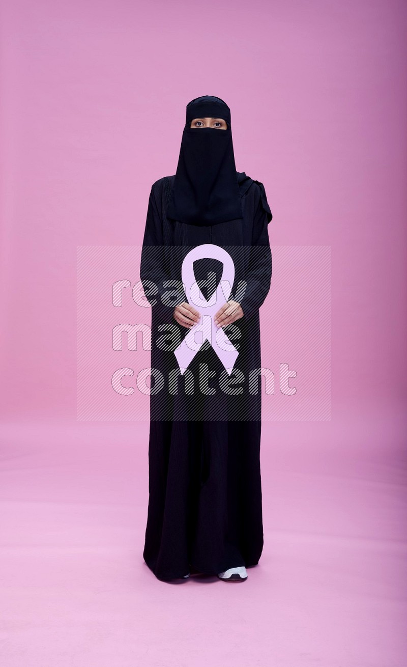 Saudi woman wearing abaya and niqab standing holding awareness ribbon on plain pink background