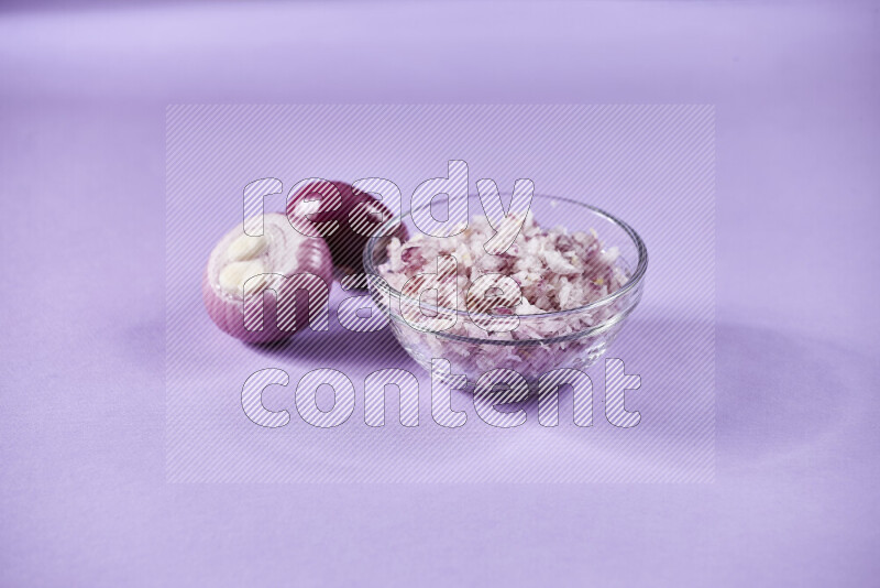 A glass bowl full of chopped red onions with halved onions beside it on purple background