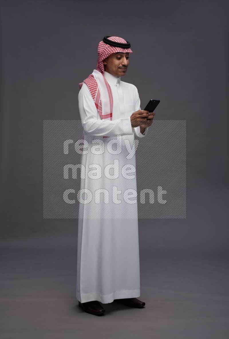 Saudi man wearing thob and shomag standing texting on phone on gray background