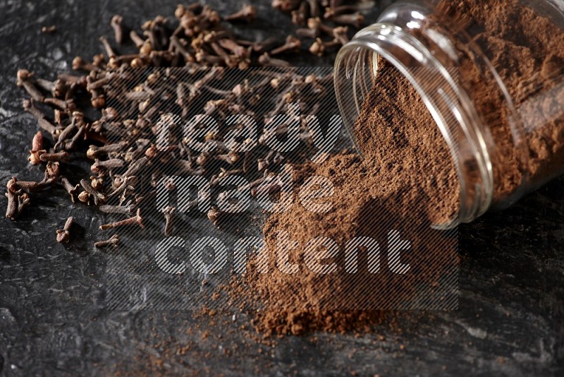 A flipped glass jar full of cloves powder with cloves spread on a textured black flooring