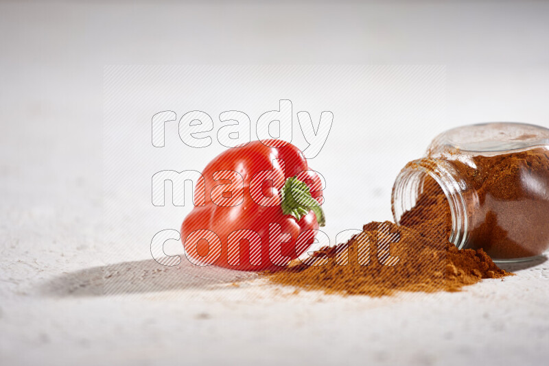 A glass jar full of ground paprika powder flipped with some spilling powder on white background