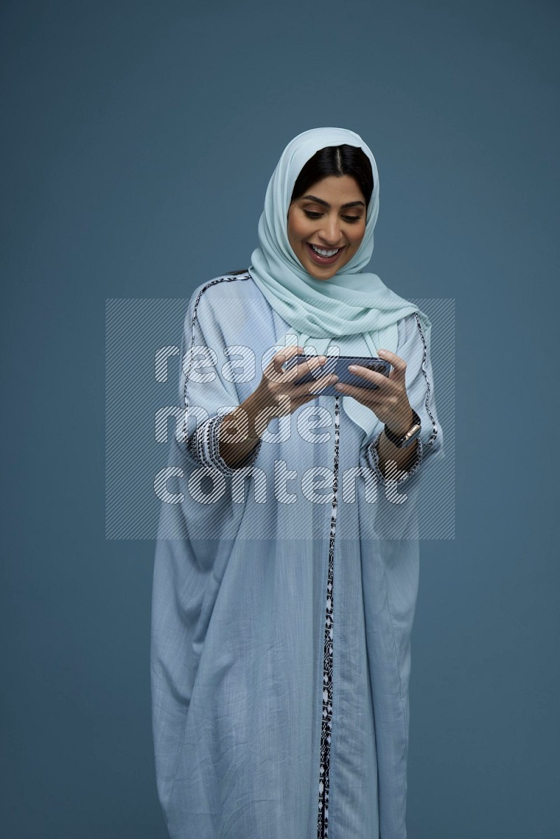 A Saudi woman Playing a Game on her phone on a blue background wearing a blue Abaya with hijab