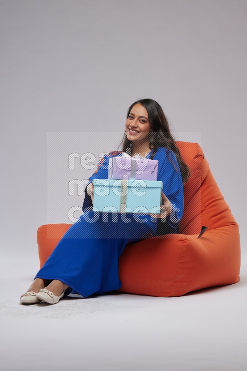 A woman sitting on an orange beanbag wearing Jalabeya holding a gift box
