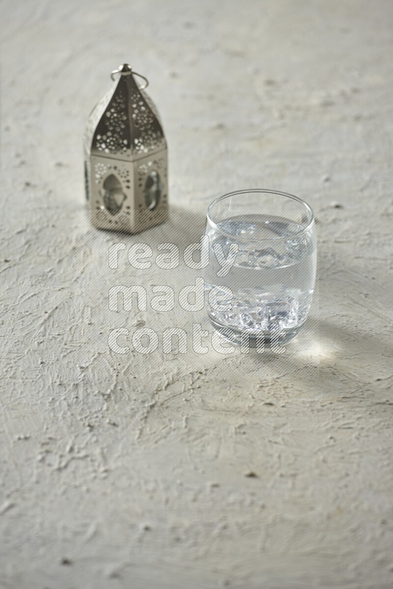 A silver lantern with different drinks, dates, nuts, prayer beads and quran on textured white background