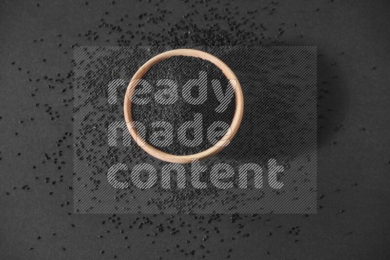 A wooden bowl full of black seeds surrounded by seeds on a black flooring