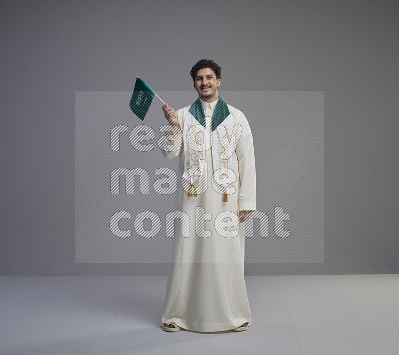 A Saudi man standing wearing thob and saudi flag scarf and holding small saudi flag on gray background