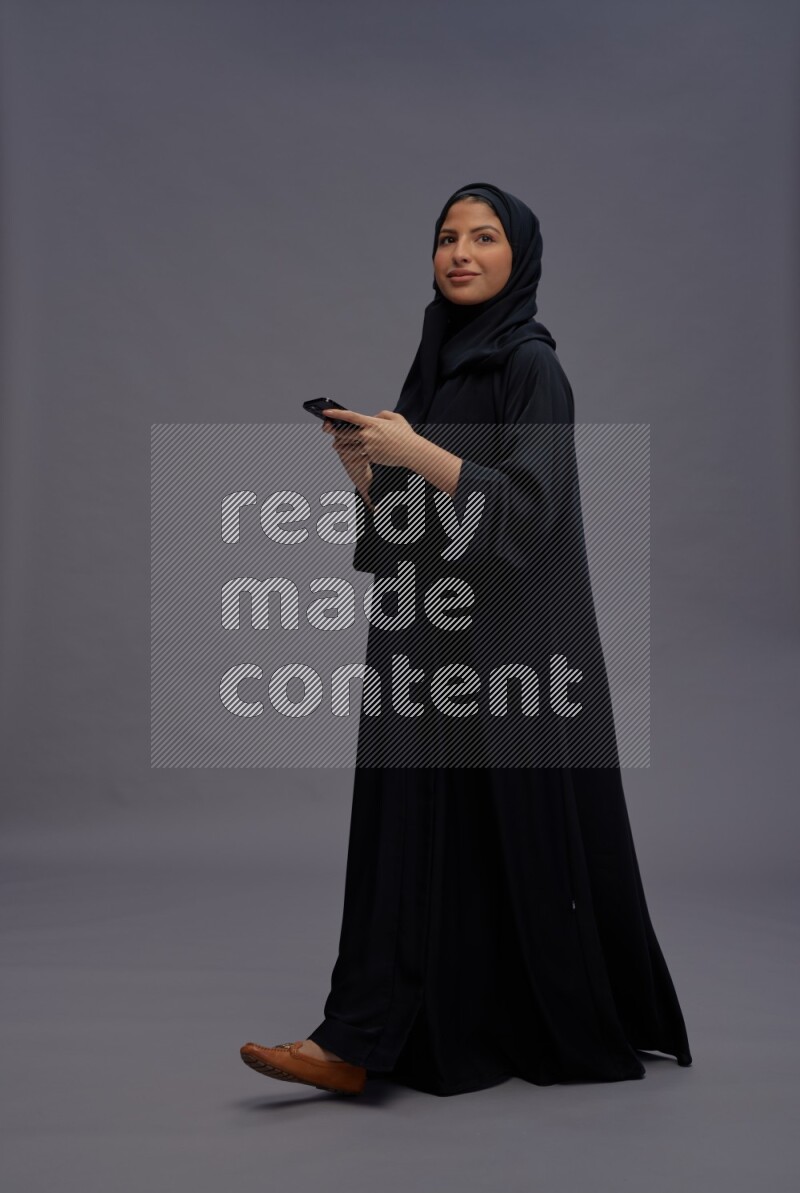 Saudi woman wearing Abaya standing texting on phone on gray background