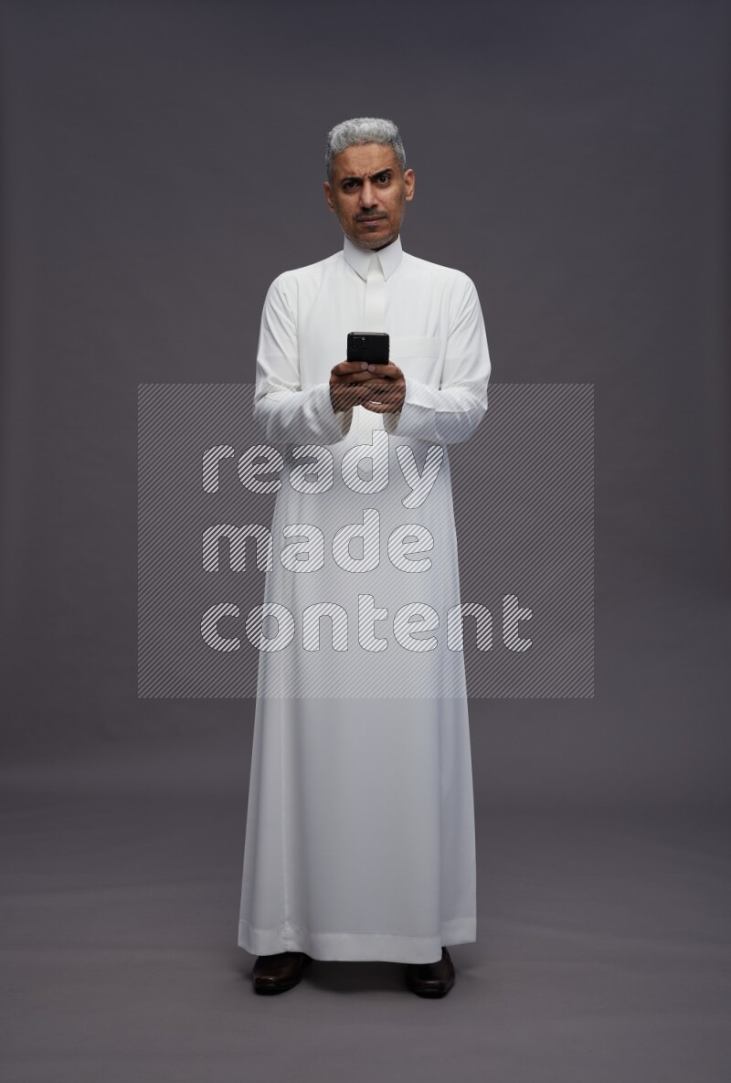 Saudi man wearing thob standing texting on phone on gray background