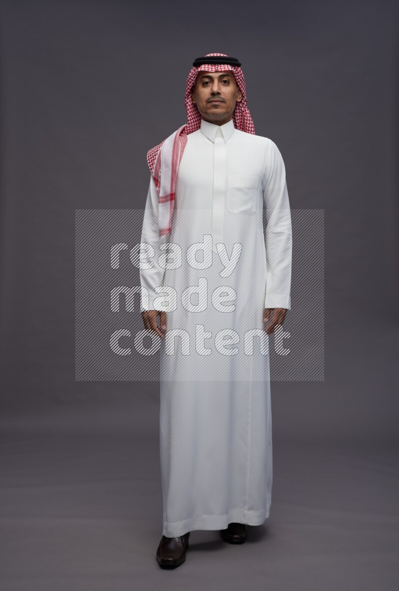 Saudi man wearing thob and shomag standing interacting with the camera on gray background