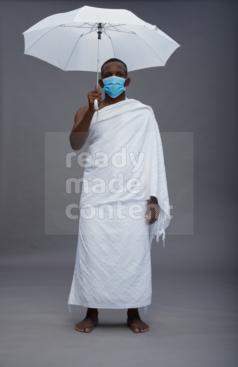 A man wearing Ehram with face mask Standing holding umbrella on gray background