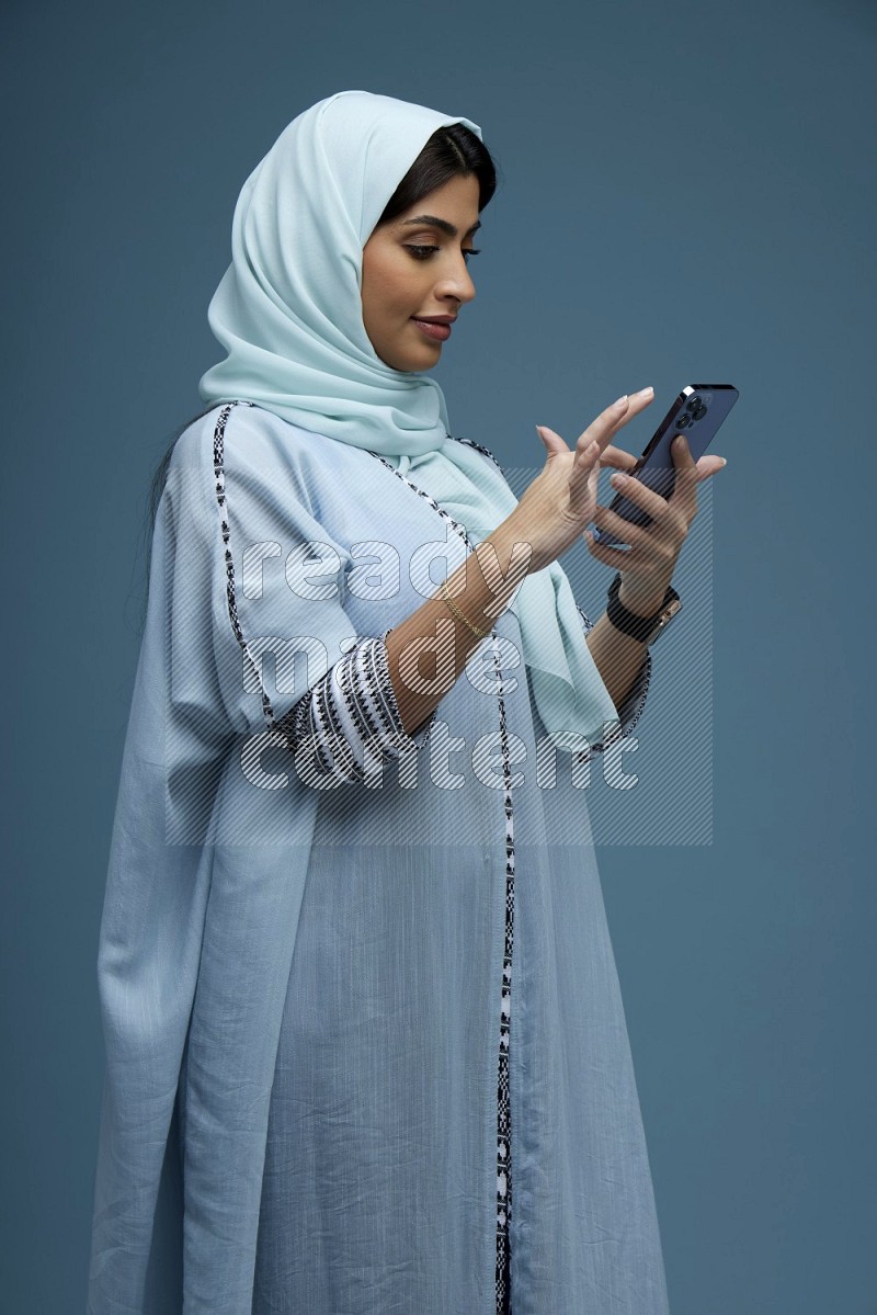 A Saudi woman Swiping on her screen on a blue background wearing blue Abaya with hijab