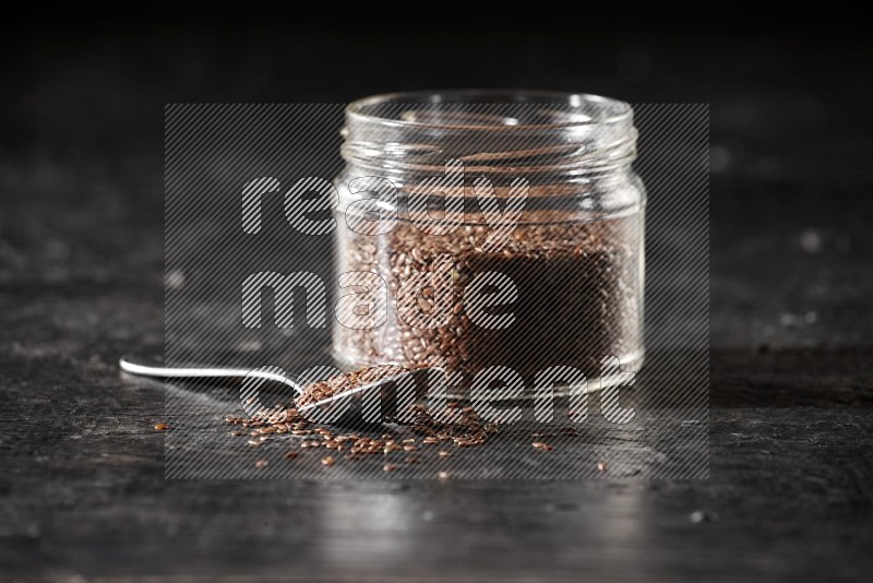 A glass jar full of flax with a metal spoon full of the seeds on a textured black flooring in different angles