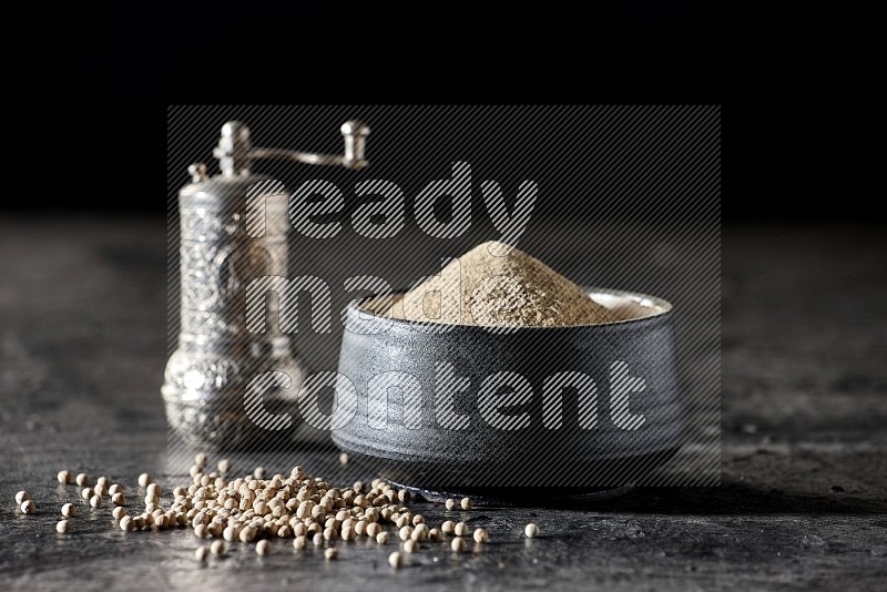 Black pottery bowl full of white pepper powder with pepper beads and metal pepper grinder on textured black flooring