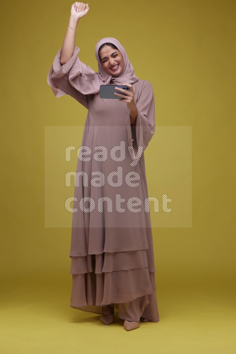 A woman Playing Games a Yellow Background wearing Brown Abaya with Hijab
