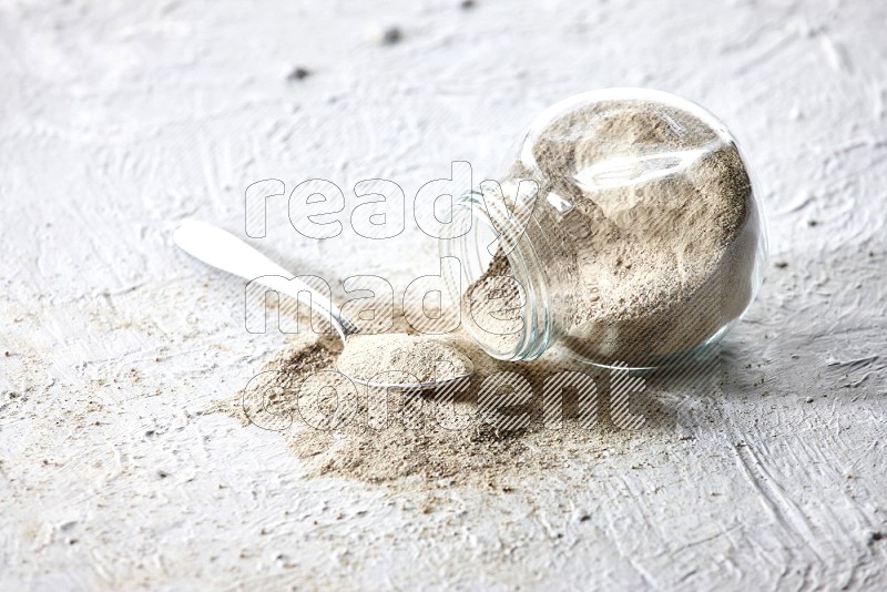 A flipped herbal glass jar and metal spoon full of white pepper powder with spilled powder on textured white flooring