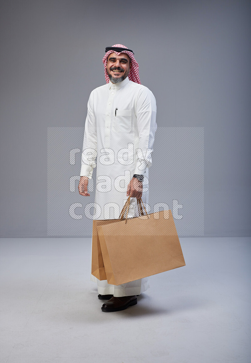 Saudi man Wearing Thob and red Shomag standing holding shopping bag on Gray background