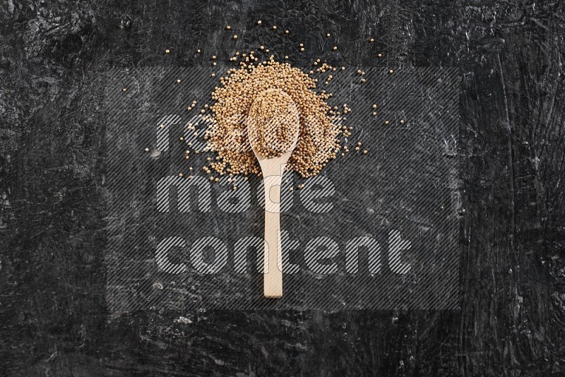 A wooden spoon full of mustard seeds on a textured black flooring