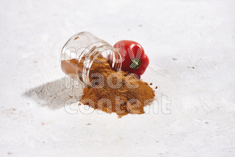 A glass jar full of ground paprika powder flipped with some spilling powder on white background